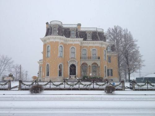 A large, ornate yellow mansion covered in snow, with a snowy landscape and trees in the background.