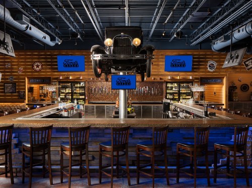 A vintage car hangs above a rustic bar with wooden stools and screens displaying the Ford's Garage logo.