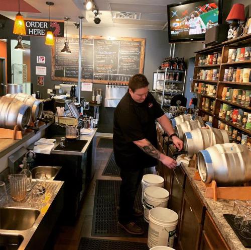 A bartender pours from a cask in a cozy bar, with shelves of drinks and a TV showing a sports game in the background.