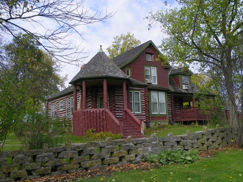 A charming red log cabin-style house with a turret, surrounded by trees and a stone wall under a cloudy sky.