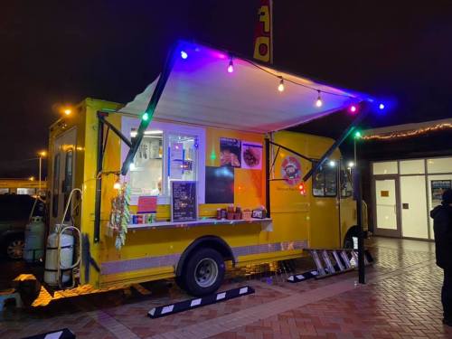 A brightly lit yellow food truck at night, decorated with colorful lights, parked in a lot.