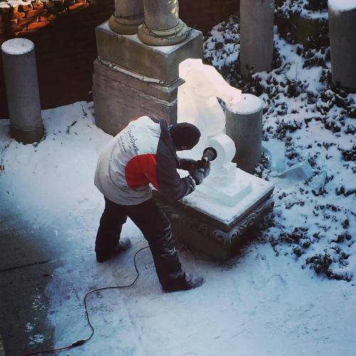 A person carves an ice sculpture outdoors, surrounded by snow and stone columns.