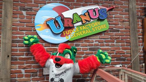 A colorful furry character poses excitedly in front of a sign for "Uranus Fudge Factory & General Store" against a brick wall.