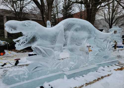 A large ice sculpture of a T-Rex surrounded by ice plants, set in a snowy outdoor environment.