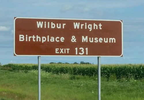 Sign for Wilbur Wright Birthplace & Museum, indicating Exit 131, with a green field in the background.