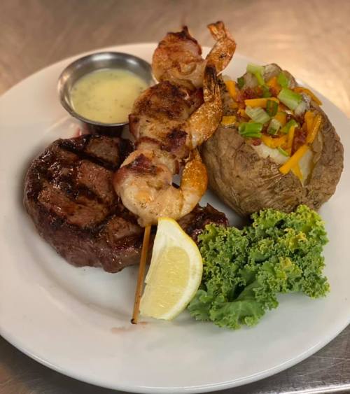 A plate featuring grilled steak, shrimp skewers, a baked potato with toppings, and a lemon wedge, garnished with lettuce.