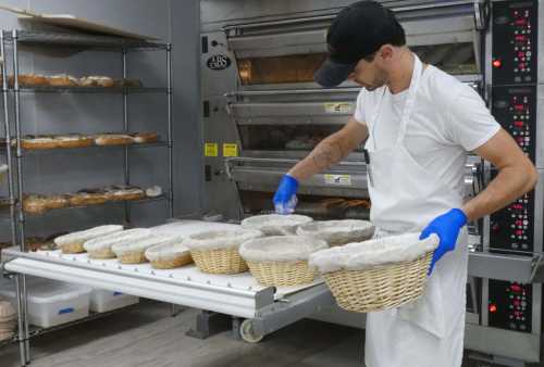 A baker in a white apron and gloves arranges dough in baskets near an industrial oven in a bakery.