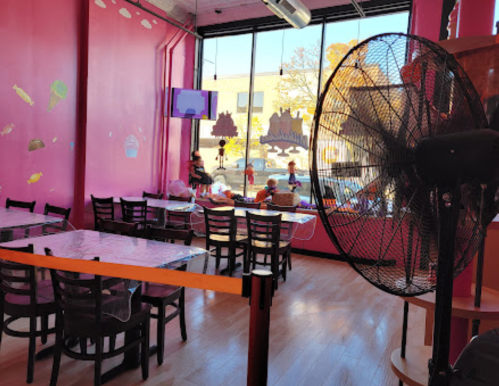A brightly colored pink ice cream shop interior with tables, a fan, and a view of people outside.