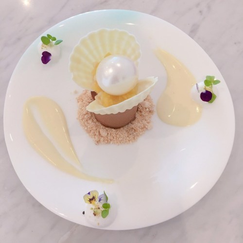 A beautifully plated dessert featuring a chocolate shell, a pearl, and decorative sauces with edible flowers.