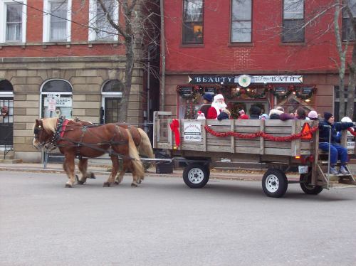 A horse-drawn wagon decorated for the holidays, with children and Santa riding through a festive town street.
