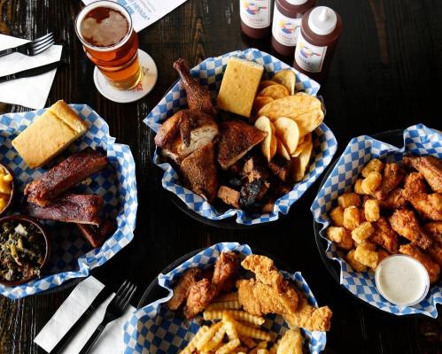 A spread of barbecue dishes including ribs, chicken, cornbread, fries, and sides, served in checkered baskets.