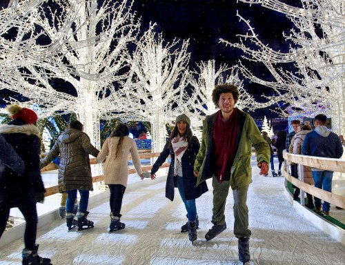 A festive ice skating scene with people skating among illuminated trees at night.