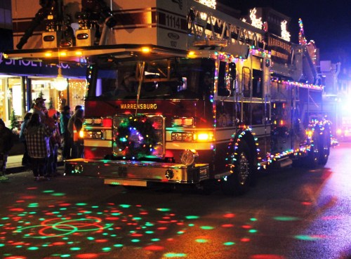 A decorated fire truck with colorful lights drives down a festive street at night, surrounded by holiday decorations.