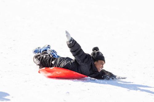 A child in a black coat and hat sleds down a snowy hill on a red sled, arms outstretched in excitement.