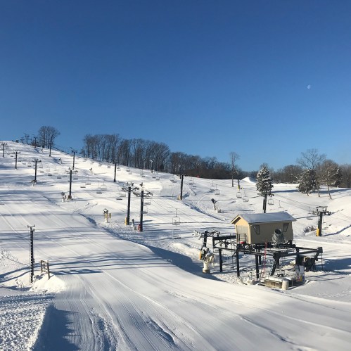 A snowy ski slope under a clear blue sky, featuring ski lifts and a small lodge in the background.