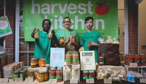 Three people stand behind a table filled with jars of food and snacks, with a green "Harvest Kitchen" banner in the background.