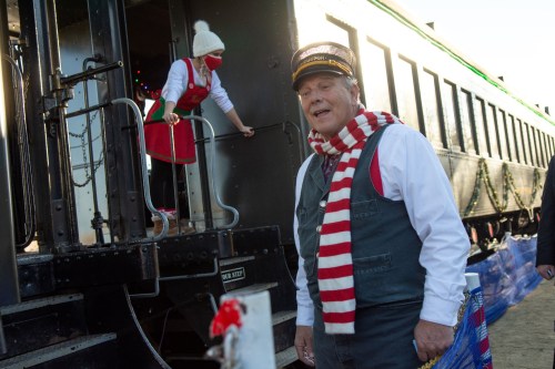 A conductor in a striped scarf stands near a train, while a woman in an apron climbs aboard. Holiday decorations are visible.