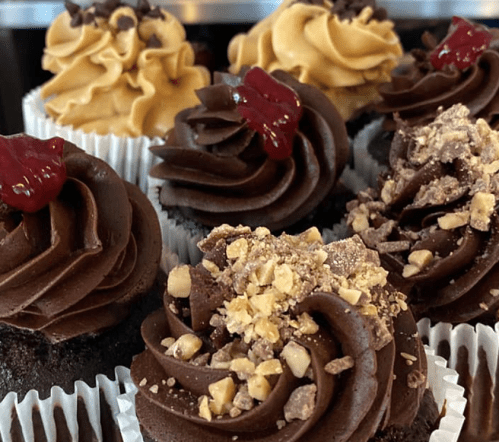 A close-up of assorted cupcakes with chocolate and yellow frosting, topped with sprinkles, jelly, and crushed nuts.