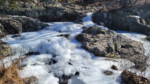 A frozen stream flows over rocky terrain, with patches of ice and frothy bubbles among the stones.