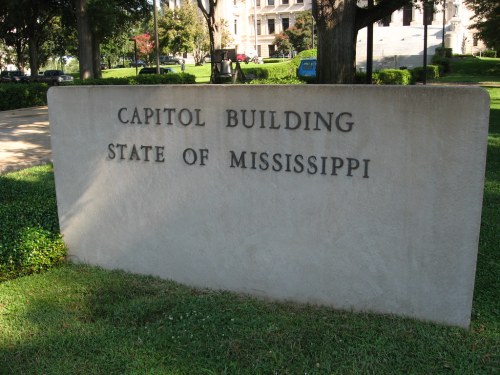 Sign reading "Capitol Building State of Mississippi" in front of a grassy area with trees.