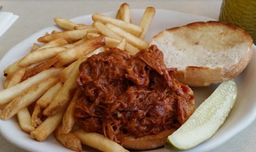 A plate with pulled pork on a bun, served with French fries and a pickle slice.