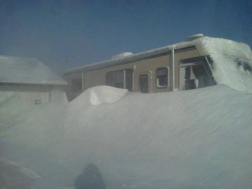 A trailer partially buried in deep snow, with a clear blue sky in the background.