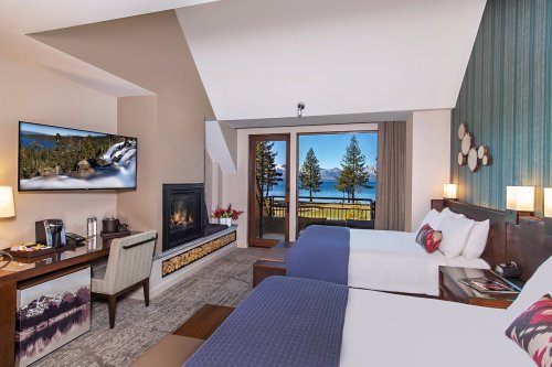 A cozy hotel room with two beds, a fireplace, a desk, and a view of a lake and trees through large windows.