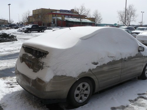 A car covered in snow and ice parked in a lot, with a restaurant visible in the background.