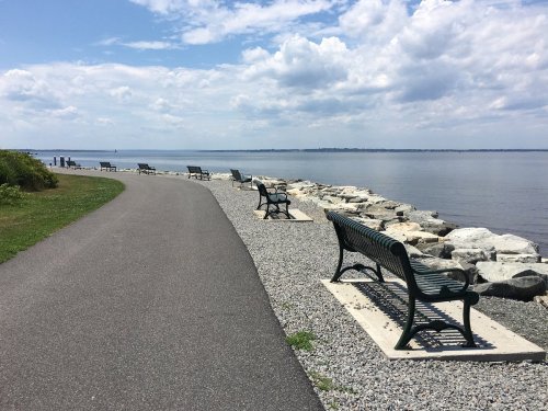 A scenic waterfront path lined with benches and rocks, under a partly cloudy sky.