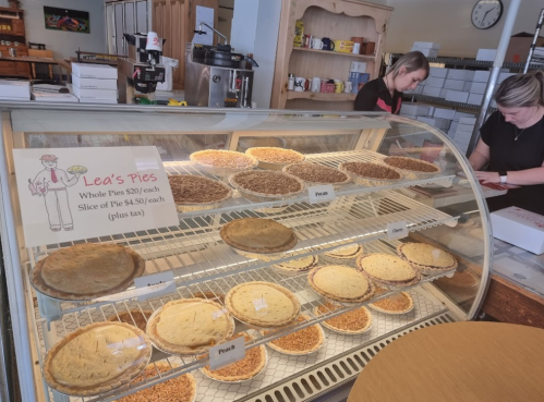 Display case filled with various pies, labeled "Lea's Pies," with whole pies and slices for sale. Two staff members in the background.
