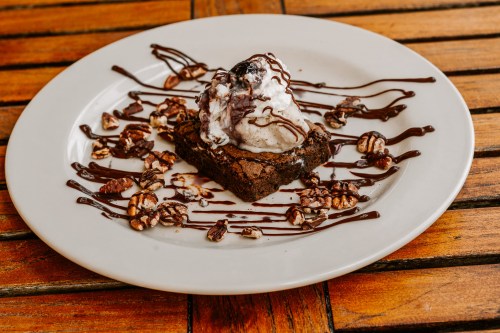 A decadent brownie topped with ice cream, drizzled with chocolate sauce and sprinkled with nuts, served on a white plate.