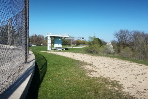 A grassy area with a sandy patch, featuring a small structure and a chain-link fence under a clear blue sky.