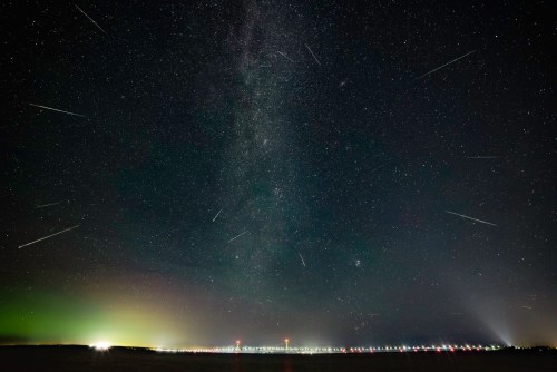 A starry night sky filled with meteors, the Milky Way visible above a distant city skyline.