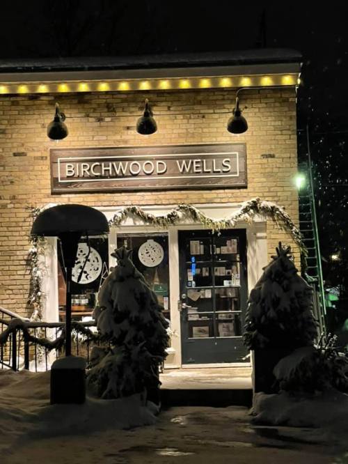A cozy brick building at night, adorned with lights and greenery, featuring a sign that reads "Birchwood Wells."