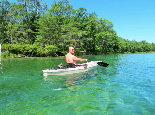 A shirtless man paddles a kayak on a clear, green lake surrounded by lush trees under a bright blue sky.