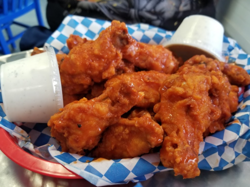 A basket of spicy chicken wings with two cups of dipping sauce on a blue and white checkered paper.