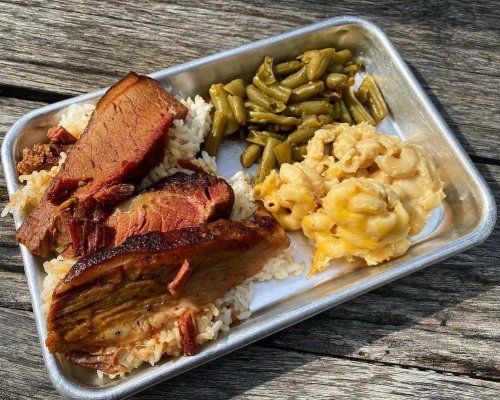 A metal tray with sliced brisket, rice, green beans, and macaroni and cheese on a wooden surface.