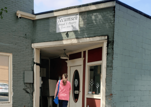 A woman walks towards the entrance of Tecumseh Bread & Pastry, a small bakery with a green exterior.