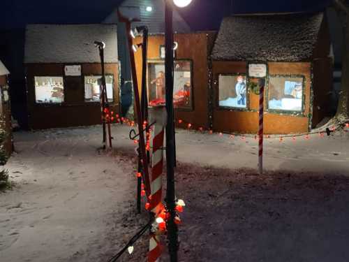 Snow-covered scene at night featuring cozy, decorated wooden houses with festive lights and a snowy pathway.