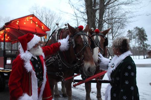 Two people in festive attire interact with two horses adorned with holiday decorations near a decorated wagon.