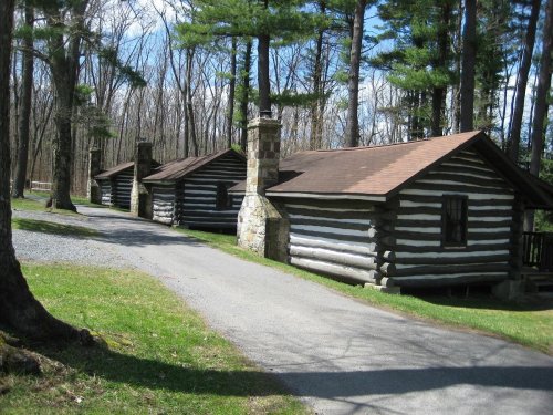 Two log cabins with stone chimneys beside a gravel path, surrounded by tall trees and a clear blue sky.