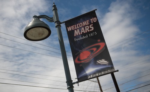 A street banner reading "Welcome to Mars" with a graphic of planets, hanging from a lamp post against a cloudy sky.
