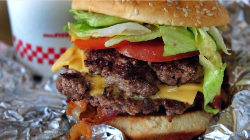 A close-up of a juicy cheeseburger with lettuce, tomato, and bacon, served with a milkshake in the background.