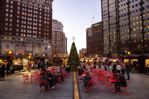 A festive outdoor market with a Christmas tree, red chairs, and holiday decorations in an urban setting at dusk.