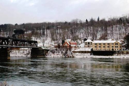 A snowy landscape featuring a river, a bridge, and houses along the riverbank, with trees in the background.