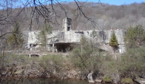 Abandoned concrete structure on a hillside, surrounded by trees and a river, under a clear blue sky.
