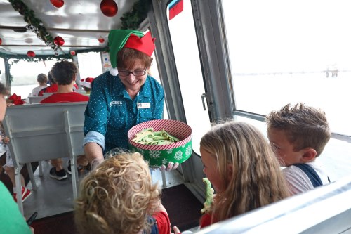 A smiling woman in an elf hat serves treats to children on a festive boat decorated for the holidays.