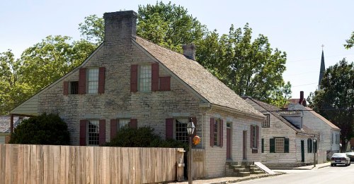 Historic stone house with a sloped roof, surrounded by trees and a wooden fence, on a sunny street.
