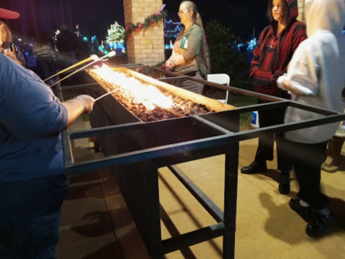 People gathered around a long fire pit, roasting marshmallows, with festive lights in the background.