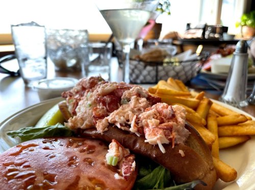 A lobster roll topped with lobster salad, served with fries, a pickle, and a cocktail in the background.